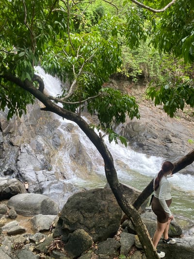 A young woman in a white long-sleeved shirt and brown shorts is sitting on a tree branch, leaning back and looking at a waterfall in Thai Mueang, Thailand. She is wearing black and white shoes. The waterfall cascades down a rocky slope, surrounded by green trees and foliage. The tree branch is angled across the top right of the scene, with the woman positioned on it. Large rocks are to the right of the branch, near the water.