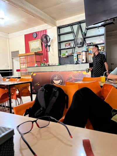 The image shows the interior of a cafe in Kutalhoksukon, Indonesia, during daylight hours. The cafe has orange plastic chairs and tables, with a counter area in the background. A man in a black and white polka dot shirt is standing behind the counter, and a person wearing black pants and a black backpack is seated at a table in the foreground, with a pair of glasses on the table. There are also drinks and food items visible on the counter and tables. The cafe appears to be moderately busy, with several tables occupied.