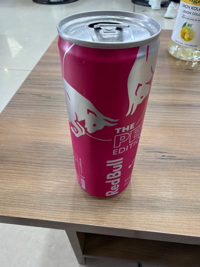 A partially consumed can of Red Bull The Pink Edition lies on a wooden surface. The can is predominantly pink with silver bull logos and lettering. The pull tab is open, and the can appears slightly dented. In the background, partially visible on the left, is a bottle of light yellow liquid. The wooden surface has a light brown, linear grain pattern. The scene does not provide any information about people, actions, interactions, or the specific location beyond the provided image. The city of Dalgopol, Bulgaria is not identifiable from the photo.