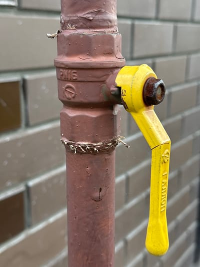 The image shows a close-up of a gas valve on a pipe. The valve is made of yellow metal and has a hexagonal bolt on top. The pipe is brown and appears to be made of metal. There is some peeling paint on the pipe, and a piece of tape is wrapped around it. The background is blurred but appears to be a tiled surface. The lighting suggests it is daytime. There is text on the yellow handle of the valve that reads "GALLON" in embossed letters. There is also a circular symbol with a line through it on the pipe. The overall impression is that of industrial equipment. There are no people in the image.