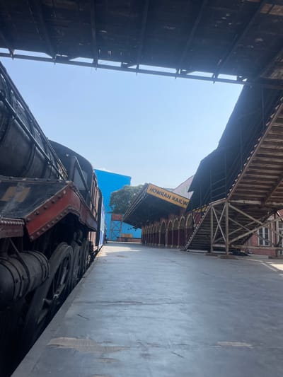 The image depicts the Howrah Railway Station in Anajpur, India. It is oriented vertically, showing the station platform, an overhead shelter, and a portion of a steam locomotive. The platform is concrete, and the shelter consists of metal beams and a roof structure. The locomotive is black with red accents and has visible wheels and mechanical components. The station signage, "HOWRAH RAILW," is partially visible in yellow lettering. The sky is blue and mostly clear. The overall environment is industrial, typical of a railway station. No people are visible in the frame.