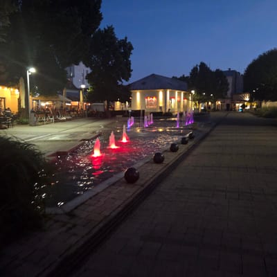 The image depicts a public square at dusk or night in the city of Piešťany, Slovakia. A fountain with colorful, illuminated water jets is a prominent feature in the foreground, with red and purple lights casting reflections on the water and surrounding paving stones. Several dark, spherical bollards are placed along the edge of the fountain area. In the middle ground, a building with a pitched roof and illuminated windows, likely a restaurant or cafe, is visible, with a sign that reads "NOVÁ KOLEKCIA". Umbrellas and outdoor seating are set up in front of establishments on the left side of the square, suggesting a commercial or dining area. Tall trees with dark foliage line the edges of the square, and streetlights are visible in the background. The overall scene conveys a lively yet peaceful urban atmosphere.