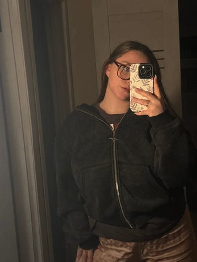 A female subject is depicted from the waist up, captured in a mirror selfie taken indoors. The scene is situated in the city of Prikubanskiy, Russia. The subject has long, straight brown hair and wears black-framed eyeglasses. Her left hand holds a smartphone with a botanical-patterned case, partially obscuring the lower right side of her face. A metallic ring is visible on her left ring finger. She is wearing a dark, faded-appearance zip-up hooded sweatshirt with a silver zipper and a star-shaped pull tab. Underneath, a dark brown t-shirt is visible. Her lower torso is clothed in light-colored, possibly champagne-hued, textured velvet or velour pants. The background shows a dimly lit interior space with a vertical light-colored trim element on the left, indicative of a wall or door frame.