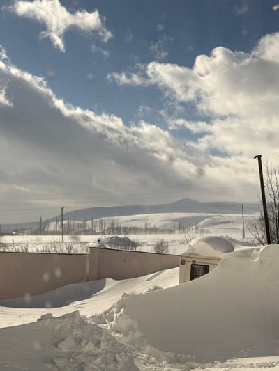 A landscape picture, tilted to the right, shows a snow-covered vista under a partly cloudy sky. White and gray clouds dominate the left side, contrasting with patches of blue. Distant, snow-dusted hills stretch across the horizon. Utility poles and bare trees sparsely populate the middle ground. Close to the camera, a structure covered in thick snowdrifts occupies the right side of the frame.