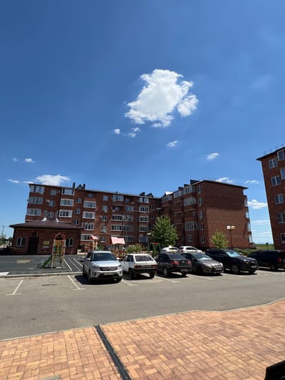 An exterior daytime, eye-level shot captures a modern apartment complex in Novaya Adygeya, Russia. The building is constructed of red brick, featuring multiple floors with numerous windows and air conditioning units. A small, attached brick structure with a brown roof is located to the left of the main building, in front of a children's playground. A parking lot is situated in front of the building, with several cars parked in designated spaces. The ground in the foreground is paved with reddish-brown bricks. The sky is a clear blue with a few scattered white clouds.