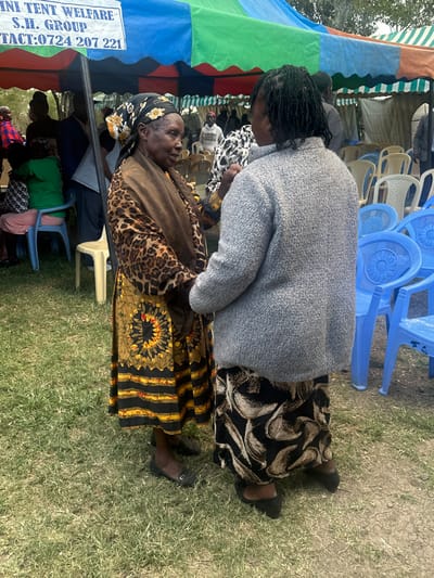 An outdoor gathering in Kiganjo, Kenya, is depicted, with individuals congregated under a multi-colored tent canopy featuring patches of blue, green, orange, and red. In the immediate foreground, two women are interacting. One woman, with dark braided hair, is seen from behind, wearing a light grey textured sweater and a black and white patterned skirt. She is leaning slightly towards an older woman, who is adorned in a patterned headscarf, a brown top with leopard print accents, and a vibrant long skirt featuring black and yellow stripes with a distinct circular, sun-like motif. Both women are wearing dark, closed-toe shoes.

The setting includes green grass covering the ground, and in the background, several stacked blue plastic chairs are prominent, alongside some cream-colored chairs. Other people are partially visible, seated or standing further back under the tent. A white banner attached to the tent's edge displays the text: "MINI TENT WELFARE S.H. GROUP CONTACT:0724 207 221". The scene suggests a daytime social or community event, characterized by its informal outdoor setup and the interaction between participants.