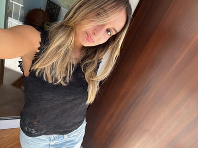 A female subject is depicted indoors, taking a selfie from a slightly low, angled perspective. The woman, appearing to be in her 20s or 30s, has long, wavy blonde and light brown hair that covers part of her forehead and left side of her face. She wears a black sleeveless top with a detailed lace overlay and scalloped trim along the armholes, paired with light-wash denim jeans. Her expression is a subtle pout, with her eyes focused directly on the camera. The background to her left features a brightly lit indoor area with a white or light-colored frame, possibly a door or window, and indistinct household objects including a dark woven basket and a framed artwork. To her right, a significant portion of the frame is filled by a uniformly dark brown wooden wall or large door composed of vertical panels. The lighting suggests natural light entering from the left. The scene is identified as being in the city of Oliveira de Frades, Portugal.