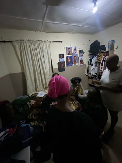 The image depicts an indoor scene, likely a room, with several individuals gathered. The room appears to be dimly lit, with a single lightbulb hanging from the ceiling providing illumination. On the wall, there are several framed photographs of people. A curtain hangs on one side of the room, and clothing items are visible on a rack in the background. The floor is covered with a patterned blanket, and some personal belongings are scattered around. The individuals are seated or standing, and their interactions are not clearly discernible due to the low light conditions and the angle of the photograph. There is no visible indication of the city of Durimi, Nigeria, within the image itself.