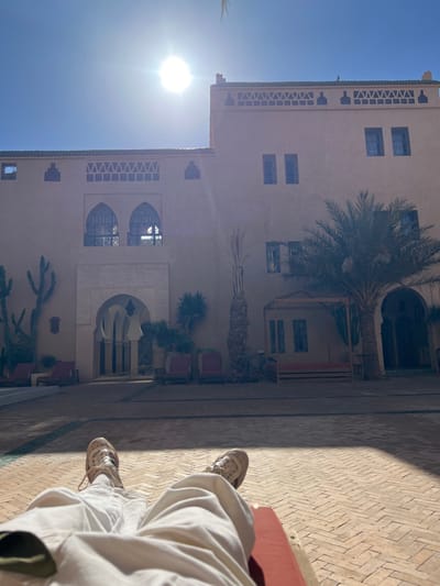 A low-angle, first-person perspective image depicts the lower body of an individual resting in an outdoor courtyard in Et-taous ⵟⴰⵡⵙ الطاوس, Morocco. The subject's legs, clad in loose-fitting light beige or white trousers and light-colored sneakers with prominent laces, extend forward, resting on a red surface, likely a lounge chair or mat, which is partially visible in the bottom right corner.
The foreground consists of a meticulously laid brick courtyard, featuring light brown bricks arranged in a herringbone pattern with darker grout lines. Straight dark lines delineate sections of the paving.
In the midground and background, a large, multi-story building constructed from earth-toned material (likely stucco or adobe) dominates the scene. The architecture exhibits traditional Moroccan influences, characterized by arched windows with intricate metal grilles, arched doorways, and decorative friezes along the roofline. A prominent central entrance features a large arched portal with multiple receding arches. Several rectangular windows are visible across the facade.
Sparse vegetation, indicative of an arid climate, is present. A tall cactus with multiple arms stands to the far left. Several palm trees, varying in height and density, are scattered across the courtyard, with one large palm tree positioned to the right of the central axis. Other smaller green plants are visible near the building's base.
Outdoor furniture is arranged around the courtyard, including several red lounge chairs, two visible to the left of the central archway and two to the right. A wooden cabana-style structure with a low bench is situated to the right, under the large palm tree.
The sky is a clear, vibrant blue, with a bright, intense sun positioned high in the upper center, casting strong shadows from the building and vegetation towards the viewer, indicating midday or early afternoon conditions.