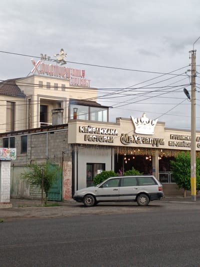 This media file shows a view of a restaurant called "Mr. Khachapuri" in Taraz, Kazakhstan. The restaurant has a distinctive sign featuring a stylized crown and the name of the establishment in illuminated letters. Above the main sign, a separate sign displays "Mr. Khachapuri Family Restaurant" with a cartoonish figure of a person in traditional attire holding a sword or knife.
The building itself appears to be a two-story structure with a beige facade. The lower part of the building has a more rustic appearance with brick and concrete block construction, while the upper story is more modern with large windows. A silver station wagon is parked in front of the restaurant, partially obscuring the entrance. Green bushes and trees are visible around the building.
The scene is set on a street with a paved road and sidewalk. Overhead power lines crisscross the sky, and a utility pole with a street lamp is present on the right side of the image. The sky is overcast, suggesting a cloudy day, and the overall lighting indicates it is daytime. There are no people clearly visible in the image, though faint silhouettes can be seen through the windows of the restaurant. The atmosphere appears calm and ordinary, capturing a typical street view of a commercial establishment.