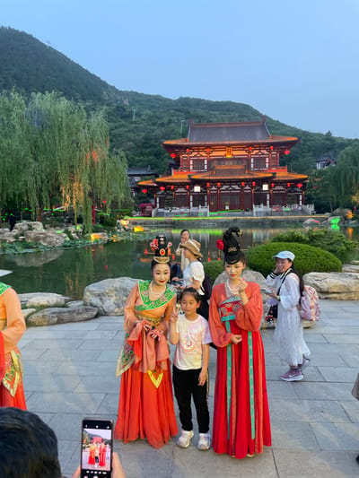 The image captures a group of people, including two women in traditional Chinese costumes, posing for a photo near a body of water in what appears to be a park or garden setting. The photo is being taken by someone using a smartphone. In the background, there is a large traditional Chinese building with red lanterns hanging, set against a green hillside. The location is Lintong District, China. The environment suggests an evening scene with the lights reflecting on the water.