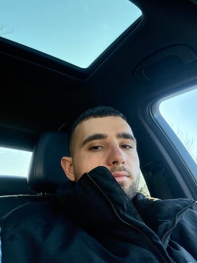 The image depicts an adult male situated within the interior of a vehicle. The male, possessing short dark hair and facial stubble, is positioned in what appears to be the front passenger seat, looking directly towards the camera. He is attired in a dark, possibly black, zippered jacket with the collar raised to obscure the lower portion of his face and neck. The vehicle's interior features a dark headliner and a visible sunroof, through which a clear blue sky is discernible. Through the vehicle's side window, bare tree branches are faintly visible against the bright sky. The specific city location of Брусовeте, Bulgaria, is not discernible from the visual content of the image.