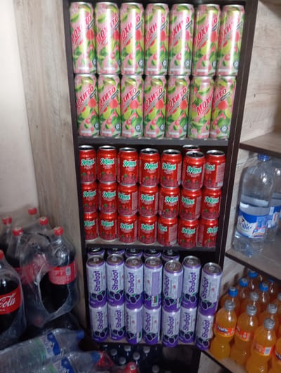 This image shows a shelf stocked with various beverages, located in Yangikurgan, Uzbekistan. The top shelf displays rows of pink and green cans labeled "Moxito Fresh." Below this, several shelves are filled with red cans bearing the "Mani" label, featuring an illustration of berries. Further down, purple cans labeled "Shaloot" are stacked. To the right of the shelves, two large plastic bottles of water are visible, one with a blue and white label and the other with a white label. In the lower right portion of the image, several bottles of orange soda, likely Fanta, are arranged. In the lower left, multiple plastic bottles of a dark carbonated beverage, appearing to be Coca-Cola, are wrapped in plastic. The environment suggests a retail setting, possibly a small shop or market.