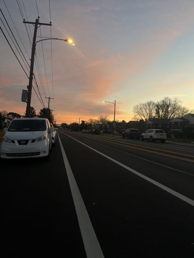 This image captures a residential street scene in Philadelphia, United States, at twilight. The sky displays a gradient of orange, pink, and light blue hues, indicating either sunrise or sunset. The perspective is from a low angle, looking down a recently paved, multi-lane asphalt road. The roadway is marked with crisp white solid lines on the edges and double solid yellow lines in the center, designating separate directions of travel.
On the immediate left, a white Nissan NV200 van is parked facing away from the viewer, with its front fascia, grille, and headlights visible. Another vehicle is partially obscured behind it. A prominent wooden utility pole on the left supports multiple power lines and a brightly illuminated street light, casting a visible flare. Further down the street on the left, another utility pole is present.
Along the right side of the road, a dark-colored SUV is parked, followed by a white Nissan Rogue SUV. Additional vehicles are parked further in the distance. A row of two-story residential houses lines the right side of the street, partially obscured by bare deciduous trees, suggesting late autumn or winter. Another illuminated street light is visible further down the road on the right. No individuals are discernible in the scene.