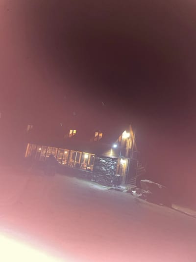 The image depicts a two-story gabled building, likely a guesthouse or restaurant, at night in Juta, Georgia. The structure features a lower facade of dark, irregular stone masonry and an upper section constructed of wood, including exposed eaves. Multiple windows on the ground floor are illuminated, revealing several indistinct human figures seated at tables inside, suggesting dining or social activity. Two smaller, rectangular windows are visible on the upper floor. An external wooden balcony with a spiral staircase descends on the right side of the building. An outdoor light fixture illuminates the area near the staircase. The ground in the immediate foreground and around the building appears covered in a light-colored surface, possibly snow or ice. Dark rock formations are visible on the right side of the foreground. The sky is dark, indicating nighttime. The overall image exhibits a reddish-pink hue, particularly prominent in the lower left quadrant.