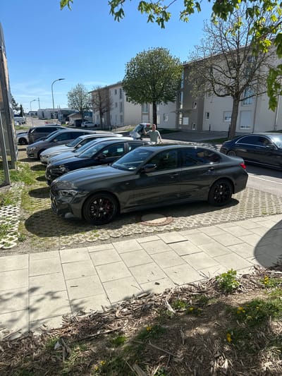 The image depicts a street scene in Jestetten, Germany. Several cars are parked in a row on the side of the road, adjacent to a paved sidewalk. A person is seen standing near the cars. Buildings are visible in the background, along with trees and a clear blue sky. The foreground shows a strip of ground with mulch and some vegetation bordering the sidewalk.