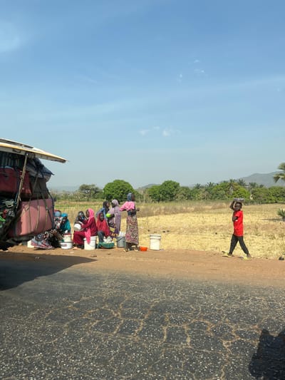 An outdoor scene in Aboro, Nigeria, depicts a roadside environment under a clear blue sky with minimal cloud cover. In the foreground, a heavily cracked asphalt road is visible, transitioning to a light brown, sandy shoulder on the right. On the far left, a white vehicle, likely a bus or large van, is partially visible, its roof rack heavily loaded with various pieces of luggage, including pink-covered items resembling mattresses or bedding, secured with ropes.
To the right of the vehicle, a group of approximately six to seven individuals, mostly women and children dressed in diverse traditional attire with head coverings, are gathered. Several are seated on the ground or low objects, while one woman in a pink top and patterned skirt stands upright. Multiple white and red buckets or containers are placed on the ground in front of the group.
Further to the right, a young boy walks from the right side of the frame towards the left, wearing a red short-sleeved shirt, dark pants, and yellow shoes. He balances a circular, flat object, consistent with a platter or bowl, on his head. The background consists of a dry, yellowish field, a line of green trees, and distant hills under a hazy sky.