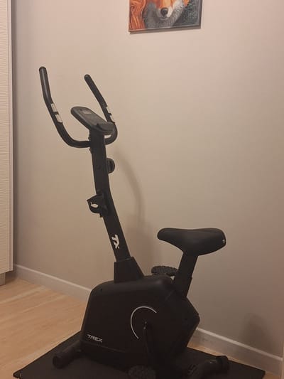 The image shows a close-up of the handlebars and console of a stationary exercise bike. The bike is black and has a digital display on the handlebars. The handlebars are curved and appear to be made of a comfortable material for grip. The console displays what looks like fitness data, though the text is too blurry to read. The word "TREX" is visible on the body of the bike. The background is a plain, light-colored wall, with a glimpse of a framed picture on the left side of the image. The overall scene suggests the exercise bike is located indoors, likely in a home or gym setting. There are no people visible in the image, and the focus is entirely on the equipment. The lighting appears to be artificial, with no indication of natural light or weather conditions. The image is taken from a slightly elevated angle, looking down at the handlebars and console. The visible text on the bike is "TX" and "TREX". The context provided indicates the location is Mińsk Mazowiecki, Poland.