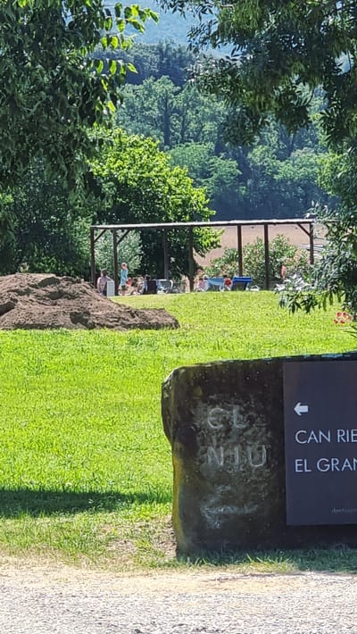 In Montagut i Oix, Spain, an outdoor scene unfolds with a focus on a grassy area and a shaded pavilion. A large mound of dark soil is prominent on the left side of the frame. Several individuals, including adults and children, are gathered under a wooden structure with a brown roof, some sitting on chairs and others appearing to be interacting. The ground is covered in lush green grass, with some patches of dry earth. In the foreground, a rough-hewn stone stands to the right, bearing carved inscriptions. A dark sign is affixed to this stone, featuring a white arrow pointing left and text in white lettering, partially visible as "CAN RIE" and "EL GRAN". The background is a dense forest with trees of varying shades of green, leading to a distant, hazy mountain range. Sunlight dapples through the leaves of trees in the foreground, casting shadows on the grass.