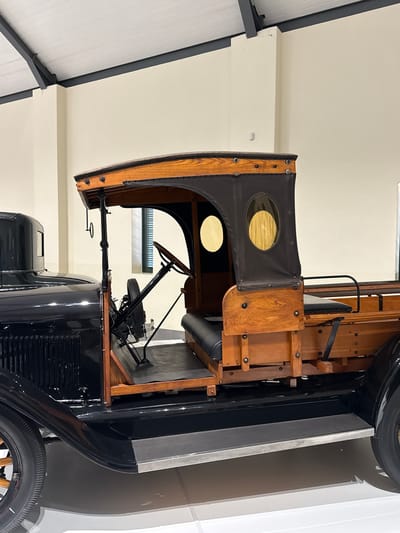 The image depicts the interior and partially exterior of a vintage vehicle, likely a truck, displayed in an indoor setting. The cabin of the vehicle is made of wood and black canvas with circular windows. Inside the cabin, a steering wheel and other vintage controls are visible. The exterior is black, with portions of the front tire visible. The vehicle is located in Stellenbosch Local Municipality, South Africa, based on visual context suggesting a museum or similar display environment. The background features a white wall.