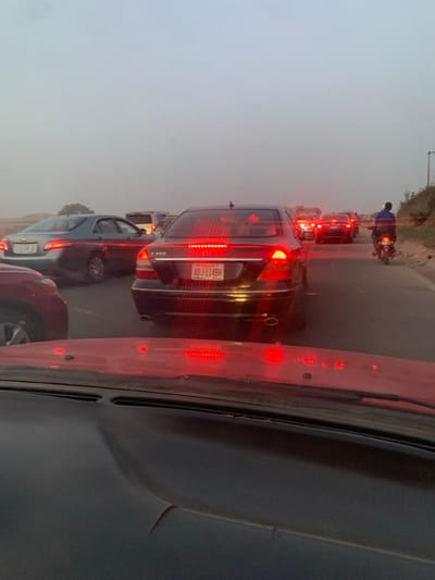 Traffic congestion is evident. Cars are lined up bumper-to-bumper on a road with poor visibility. A motorcycle weaves through the cars. The focus is on the brake lights of a black Mercedes-Benz E350 with license plate ABJ-114BH. The photo appears to be taken from inside a car, showing part of the dashboard and windshield. The overall scene conveys a sense of slow-moving or stalled traffic.