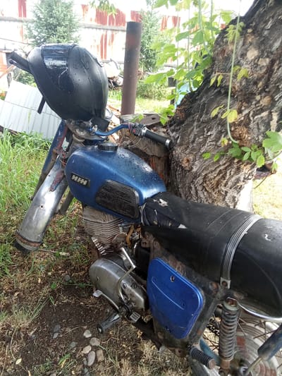 A dilapidated blue motorcycle is parked outdoors, leaning against a large tree in a rural setting. The motorcycle appears to be old and weathered, with visible rust and wear on its parts. The front fender is detached and leaning against the ground. The seat is black and appears worn. The motorcycle is covered in water droplets, suggesting recent rain. The background shows a rustic fence, some evergreen trees, and overgrown grass and weeds, consistent with a rural or semi-abandoned outdoor area in Бородулиха ауылдық округі, Kazakhstan. There are no people visible in the image.
