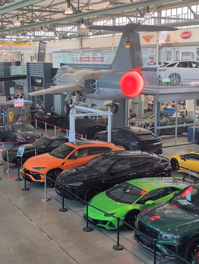 This media file captures a view of a museum or exhibition hall filled with various vehicles, including cars and an airplane. The setting appears to be indoors, with exposed metal beams and lighting fixtures visible in the ceiling, suggesting an industrial or large exhibition space.

The main subjects are a collection of luxury sports cars, predominantly in bright colors like green and orange, parked in a line and separated by stanchions. Among these is a striking green Bentley and an orange Lamborghini. An airplane, likely a military transport or cargo plane, is prominently displayed with its propeller visible, angled downwards and to the left. It features a German flag emblem on its tail.

There are a few people visible, mostly in the background and appearing to be visitors or staff members, some are walking and some are standing. They are not the primary focus of the image. The scene seems to be a public exhibition of vehicles.

Notable details include signage, such as a "BUGATTI" oval logo high on a wall in the background, and other signs that are difficult to read clearly. The lighting suggests it is daytime, but the interior setting makes specific time of day hard to determine. There are no clear indications of weather conditions. The overall impression is of a well-lit, organized exhibition space showcasing high-performance vehicles and an aircraft.

Visible text includes: "BUGATTI", "MOTORWORLD", "LIQUI MOLY", and "Solitude". The German flag colors are also visible on the airplane.