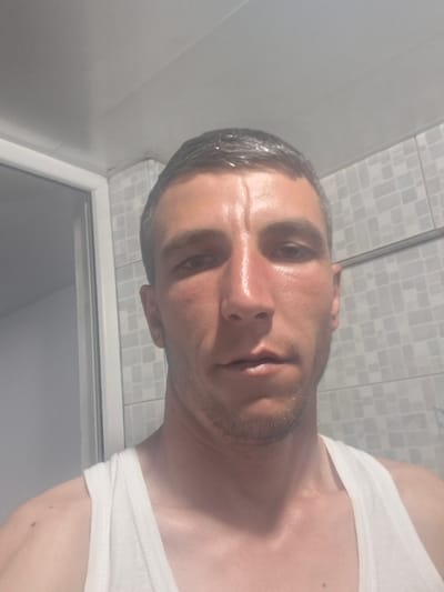 The image is a selfie of a light-skinned man in a white tank top, taken from the chest up. He has short, dark hair, a prominent nose, and a slight stubble. The background appears to be a bathroom, with tiled walls and a shower door visible. The lighting is bright, suggesting an indoor setting. The image does not contain any information about the city of Hovtamej, Armenia.