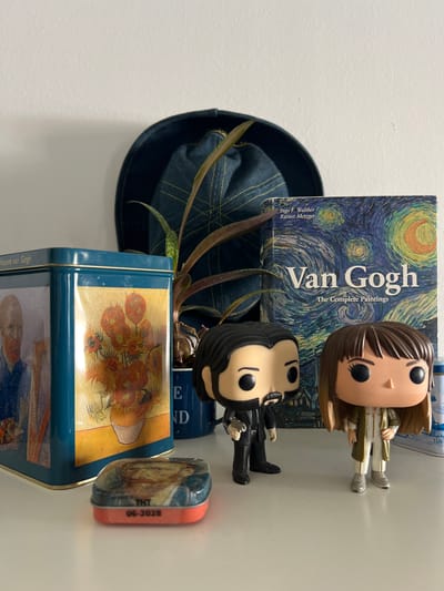 The image depicts a white shelf with several objects. On the left is a teal tin featuring Vincent van Gogh paintings, including a portrait and sunflowers. In front of the tin is a small, rectangular tin with an image of a portrait and text that reads "THT 06-2028". To the right of the tin are two Funko Pop figures: one resembling John Wick and another resembling a female character. Behind the figures is a book titled "Van Gogh: The Complete Paintings" with a depiction of "The Starry Night" on the cover. Further to the right, a portion of a blue and white patterned object is visible. Behind the objects, a dark blue denim hat and a potted plant with long, narrow leaves are placed against a white wall. The scene does not contain any information that would allow for its location to be determined as Mollet Del Vallès, Spain.
