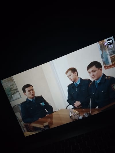 Three men in dark blue uniforms are seated at a wooden table. The man on the left is wearing a dark blue collared shirt with epaulets and a dark blue jacket with insignia. He has a lanyard with an ID card clipped to his jacket. He is looking to his right and appears to be speaking. The man in the middle is looking down and appears to be listening. He is wearing a similar uniform. The man on the right is looking forward and slightly to his left. He is also wearing a dark blue uniform with epaulets and insignia. The background is a plain, light-colored wall. There is a map on the wall behind the man on the left. On the table are two objects, one appearing to be a decorative globe and the other a small metallic object. The image is angled and the edges are blurred. There is a watermark at the bottom left of the image that reads "vk.com/multik_yar". A logo in the top right corner contains the word "КИНО". The provided coordinates of Dovzhansk, Ukraine do not align with the visual information in the image.