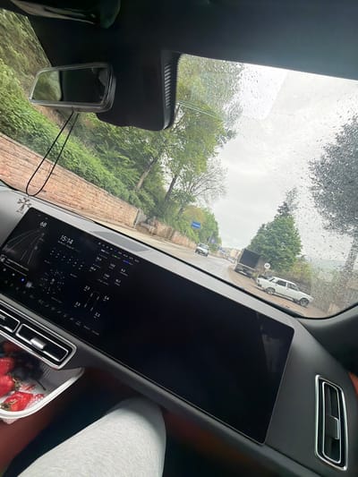 The image, taken from inside a car, shows a rainy day in Ijevan, Armenia.  The dashboard features a large touchscreen displaying various car metrics, time 15:14, date May 11, cloudy 10*C, and a music selection.  A rearview mirror is visible. Through the rain-streaked windshield, a street scene unfolds with green trees lining the road and buildings visible in the background. Cars can be seen on the road. A white car is parked on the right side of the road near a 40 km/h speed limit sign. To the right of the dashboard, part of a passenger's leg in white leggings is visible, along with a container of strawberries. A cross necklace hangs on the mirror in front of the dashboard.