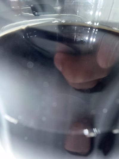 The image shows a close-up, out-of-focus view of a dark liquid in a clear container, possibly a glass or bottle. The liquid has a frothy or bubbly surface. A person's hand or finger is visible, partially submerged in the liquid and also seen reflected on the surface. The lighting creates highlights and reflections on the container and the liquid's surface. There is no discernible information in the image that can be used to identify a specific location in Buchholz in der Nordheide, Germany, or any details about a crime scene. The scene is not forensic.