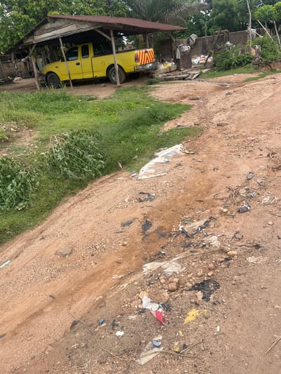 The image depicts a dirt road on the outskirts of Osogbo, Nigeria. Litter is scattered along the road. A yellow pickup truck is parked under a dilapidated shelter to the left of the road. Vegetation is growing next to the road.