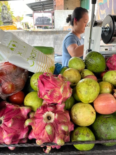 This media file captures a vibrant, close-up view of a fruit stand in Tabanan, Indonesia. The scene is dominated by an assortment of fresh produce, including bright pink dragon fruits with their characteristic spiky exteriors, numerous green and yellow guavas, and a few red tomatoes. Two avocados are also visible among the fruits.

In the background, a woman is partially visible, leaning over a machine, possibly a juicer or a food preparation device, suggesting that fruits are being processed or sold as juice. She is wearing a casual blue t-shirt, and her hair is tied back.

A white plastic bag with green and yellow striped accents is prominent in the foreground. Visible text on the bag indicates it is for "disposable hand gloves" and lists various uses such as "Home used," "Hygiene," "Salon used," "Catering used," "Medical used," and "Chemical used." A small fly is perched on the bag, a detail that adds to the authenticity of the outdoor market setting.

The lighting suggests it is daytime, and the overall impression is one of a bustling local market scene, alive with the colors and freshness of tropical fruits. The arrangement of the fruits and the presence of the processing equipment point towards a small business operation, likely a roadside stall or a stall within a larger market. The focus is on the abundance and variety of the fresh produce available.