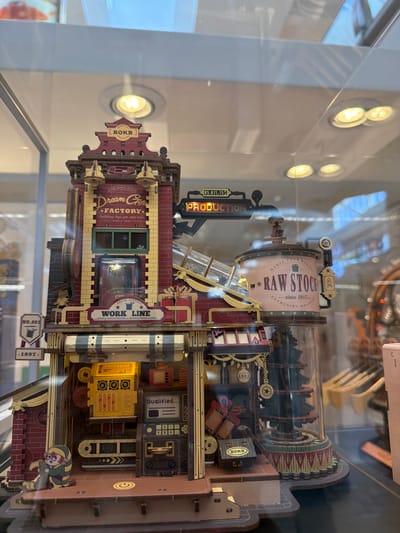 The image shows a detailed model of a factory, possibly a toy factory, displayed behind glass. The factory is multi-storied and made of wood, with intricate details and signage. Prominent labels include "ROKR," "Dream Glow FACTORY," "PRODUCTION," "RAW STOCK," and "WORK LINE." There are miniature figures, machinery, conveyor belts, and decorative elements like gift boxes and a Christmas tree inside a glass dome. The overall aesthetic suggests a whimsical and vintage theme. The model appears to be part of a retail display, as indicated by the reflections of overhead lighting and display cases in the background. The location is Mission Viejo, United States of America.