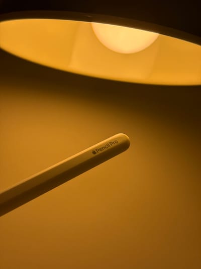 In the city of Kfar Saba, Israel, an Apple Pencil Pro is illuminated by the warm glow of a lamp, casting a soft shadow on a textured, orange-hued surface. The pencil, positioned diagonally from the bottom left to the upper right of the frame, displays its branding: a subtle Apple logo followed by "Pencil Pro" in a minimalist font. The lamp's shade dominates the upper portion of the image, its curved interior softly diffusing the light from the bulb. The overall impression is one of focused light and shadow, highlighting the sleek design of the stylus against a warm, ambient backdrop.