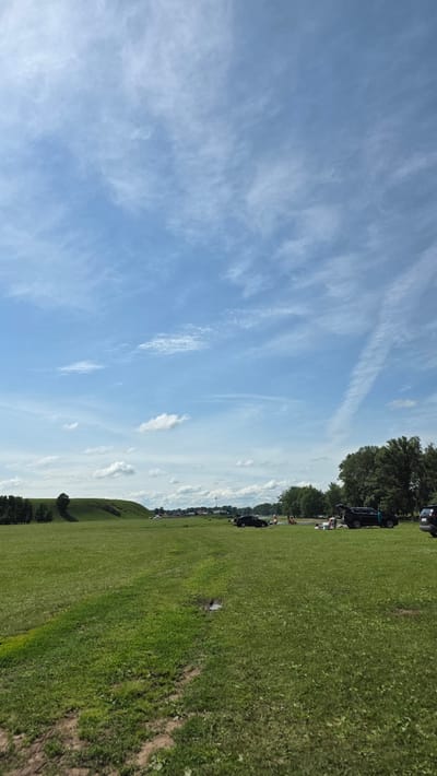 The image depicts a wide, grassy field under a bright blue sky with scattered white clouds. In the distance, a gentle, tree-lined hill is visible on the left. A line of trees forms the horizon on the right side of the frame. Several vehicles, including a black car and a dark SUV with its trunk open, are parked on the grass. A group of people are gathered near the vehicles, some appearing to be picnicking or relaxing. The overall scene suggests an outdoor recreational area, possibly a park or open space, in the vicinity of Таш-Асты, Russia.