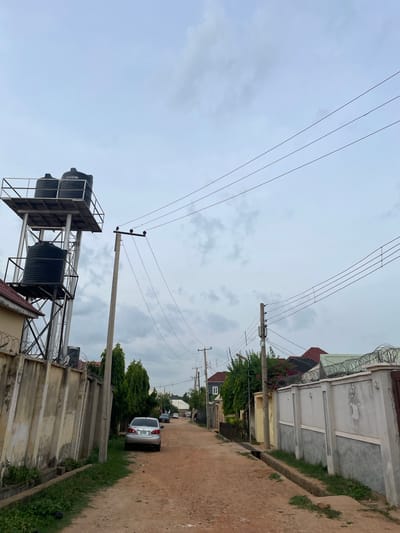 A dirt road winds through a residential area in Unguwan Dosa, Nigeria, under an overcast sky. On the left, a tall metal tower supports two large black water tanks, one above the other. Adjacent to the tower is a textured concrete wall topped with barbed wire. Further down the road, a silver sedan is parked on the right side. On the right side of the road, a beige concrete wall, also adorned with barbed wire, encloses properties. Several power lines crisscross the sky above the scene, supported by concrete and wooden utility poles. Lush green trees and some flowering bushes are interspersed among the houses, which exhibit a mix of architectural styles with pitched roofs. The ground is predominantly composed of reddish-brown earth, with patches of green grass growing along the roadside.