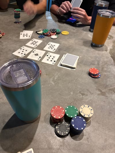 A poker game in progress on a grey surface. Cards are dealt, displaying combinations like three of a kind and straights. Multiple stacks of colorful poker chips are scattered around, indicating active betting. Players' hands are visible, holding cards. Two large insulated cups, one blue/yellow and another teal, sit nearby, presumably for drinks. One small stack of cards sits face-down.