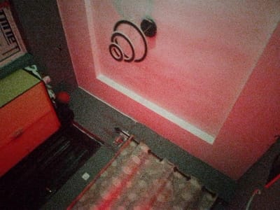The image depicts an interior room in Kongo, Nigeria, captured from a high, tilted perspective looking downwards. The ceiling and a portion of the visible wall are painted a reddish-pink color. A recessed section of the ceiling is highlighted by a white border. Mounted centrally on the pink ceiling is a black, decorative fixture composed of multiple concentric or spiraling rings. The floor is dark grey or black. A patterned rug, displaying light and dark circular patterns with a prominent red stripe, covers a segment of the floor. Small, dark, cylindrical objects, possibly pins or anchors, are visible along the rug's edge where it meets the floor. In the upper left, a piece of furniture, likely a cabinet or chest of drawers, is visible with a light green top and an orange-red body, featuring at least one dark handle. A small, spherical red object rests on the floor next to this furniture. A thin, dark, linear object is situated on the floor adjacent to the rug. No individuals are present. The image exhibits significant grain.