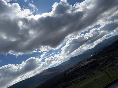 A diagonally oriented landscape photograph depicts a mountainous region in the vicinity of Real Sitio de San Ildefonso, Spain, under a dynamic sky. The upper two-thirds of the frame are filled with an extensive cloud cover consisting of large, dense, dark grey clouds intermingled with patches of bright blue sky. Direct sunlight is visible breaking through the clouds in the upper left quadrant, illuminating cloud edges and casting light upon the scene below.
In the mid-ground, a prominent mountain range extends across the view. The higher elevations of these mountains are clearly marked by white snow caps. Their slopes are covered in dark green and brown vegetation. Situated at the base or on the lower slopes of the mountains, a cluster of buildings with light-colored roofs indicates a settlement, identified as part of Real Sitio de San Ildefonso.
The lower foreground features green, compartmentalized fields, separated by what appear to be fences or property lines. An indistinct, dark structural element occupies the extreme lower right corner of the image. No individuals, specific actions, or direct human interactions are present in the scene. The environment is characterized by natural mountainous terrain, variable atmospheric conditions, and localized human habitation.