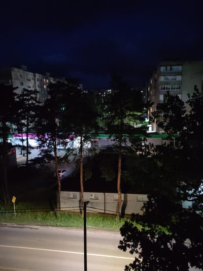 The image depicts a nighttime street view in Kotovsk, Russia. The scene is dominated by tall pine trees silhouetted against the dark sky and illuminated apartment buildings. Several cars are parked in a lot or along the street. The buildings appear to be residential, with some windows lit from within. A road with white lane markings runs in the foreground, and a grassy area with a yellow sign is visible to the left. The overall atmosphere is quiet and urban, with the artificial lights creating a stark contrast to the natural darkness.