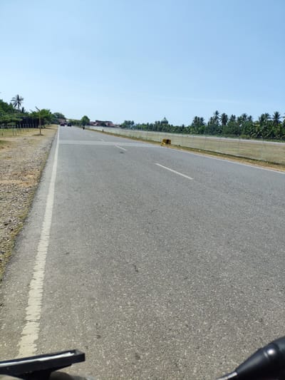 The image depicts a paved road with a white line marking its left edge. The road is a two-lane asphalt surface, with white dashed lines dividing the lanes and solid white lines marking the center of the road. There is a sandy shoulder on the left side of the road, with some sparse vegetation. In the background, there are buildings with red roofs and a green dome, possibly a mosque, visible amongst palm trees and other tropical foliage. A chain-link fence runs along the right side of the road, separating it from a grassy field. Several vehicles, including cars and a motorcycle, are visible on the road in the distance. The sky is clear and blue. The image appears to be taken from a low angle, possibly from a moving vehicle, given the presence of handlebars in the foreground. The location is identified as Keupoknibong, Indonesia.