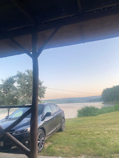A black station wagon is parked on a grassy area next to a body of water. The car is viewed from the front driver's side, with its headlights and front grille visible. The scene is framed by the wooden structure of a gazebo or porch, with posts and a roof overhead. The body of water appears to be a lake or river, with a misty haze on its surface. Trees and vegetation line the far bank of the water. The sky is a pale blue with a hint of pink or orange near the horizon, suggesting either dawn or dusk. The overall impression is of a peaceful, outdoor setting. The location is identified as Găleștii Noi, Moldova.