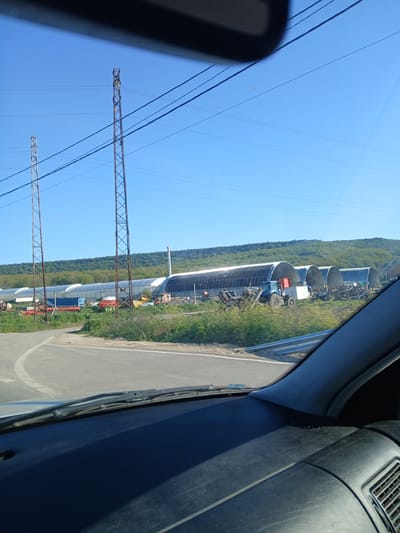 The image shows an outdoor scene from inside a vehicle, possibly a car or truck. The environment consists of a clear blue sky with visible overhead power lines supported by lattice towers. Adjacent to the road is a series of long, arched structures resembling airplane hangars or elongated greenhouses. Various pieces of farm equipment, including tractors, are parked near these structures. The terrain adjacent to the road is grassy with patches of vegetation, backed by a dense line of trees and forest. The location is possibly near Tsarkva, Bulgaria, based on the presence of agricultural structures and terrain characteristic of that region.