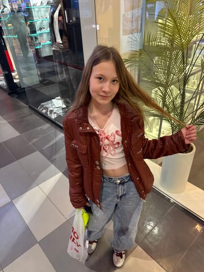 A young girl stands on a checkered tiled floor in what appears to be a shopping mall. She is wearing a brown jacket over a white top with a pink design, blue jeans, and sneakers. She is holding a strand of her long, light brown hair in her right hand. In her left hand, she is holding a white shopping bag with red lettering and a hint of a bright yellow item inside. Behind her, there is a display case with various items on shelves and a large potted plant. The image suggests an indoor environment with modern flooring and potentially commercial establishments nearby. While the image provides visual details, determining the specific location in Ol'ginskaya, Russia, is not possible without additional contextual information.