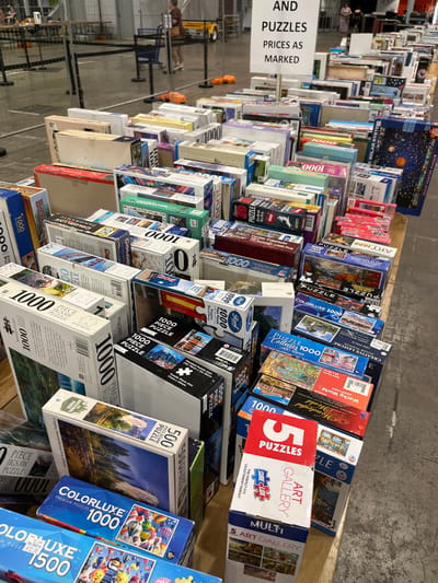 The image displays a large collection of jigsaw puzzles arranged for sale in an indoor market or retail setting, likely in South Brisbane, Australia, given the provided context. Hundreds of puzzle boxes are densely stacked and displayed on long, wooden tables, creating rows of merchandise.

The primary subjects are the numerous puzzle boxes, showcasing a wide variety of piece counts and themes. Many boxes clearly state "1000 PIECE PUZZLE" and "500 PIECE JIGSAW PUZZLE," with some "1500" piece puzzles also visible. Themes range from natural landscapes, often featuring "NATIONAL PARK" imagery, to celestial scenes like "SOLAR SYSTEM" or "GIANT POSTER PUZZLE." Other identifiable themes include "ART GALLERY" collections, "Wacky World," and "Wonderful World" puzzles, along with various abstract or scenic designs. Several brands are visible, including "COLORLUXE," "Reed," and "SURE LOX." A distinctive "5 PUZZLES" pack is also prominently displayed.

The event appears to be a sale or a section dedicated to puzzles within a larger store or market. A white sign on the left-hand side of the display is clearly legible, stating "AND PUZZLES PRICES AS MARKED," indicating that prices are individually marked on each box.

The setting features concrete flooring and metal shelving units in the background, suggesting a warehouse-like commercial space. Blurry figures of people, likely shoppers or staff, can be seen in the distant left background. The lighting is artificial, characteristic of an indoor environment, making the specific time of day unidentifiable. The overall impression is one of an extensive selection of puzzles available for purchase.