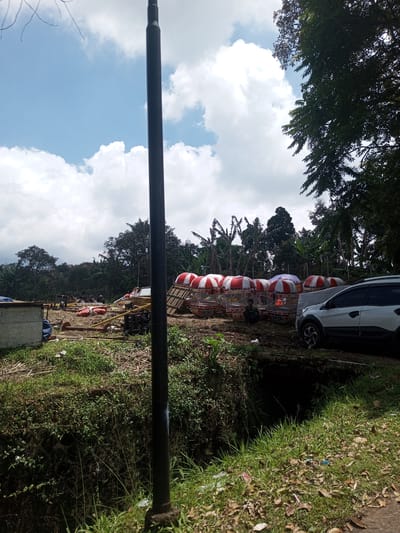 The scene is an overgrown area, possibly on the outskirts of Kioumaknao 2, Laos. A light pole dominates the foreground. Further in the background, a collection of red and white birdcages are arranged near a white SUV. A person is seated next to the cages. To the left, there is debris, possibly construction materials, scattered across the ground. Trees and other vegetation form the backdrop. There is a small ditch or depression in the ground, partially hidden by grass. The sky is blue with scattered white clouds.
