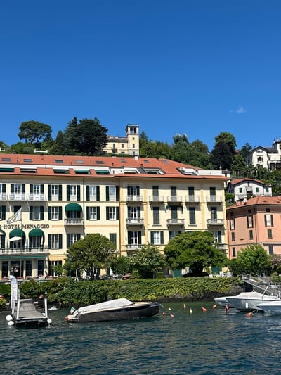 This daytime photograph presents a tranquil lakeside scene in Loveno, Italy, likely on Lake Como, bathed in bright sunlight under a clear blue sky dotted with a few wispy clouds.
In the foreground, the calm, rippling water of the lake reflects the light. Several boats are present; a large, dark-hulled boat, partially covered, is moored near the verdant shoreline, which is densely covered with green ivy and bushes. A small, modern pier extends into the water, with a lighter-colored boat tied to it. Orange and yellow buoys float in the water, and an Italian flag (green, white, red) is visible on one of the smaller boats near the pier.
The midground features a prominent, multi-story hotel building with a pale yellow facade. It boasts numerous windows with dark green shutters, balconies with ornate white railings, and several green awnings. The name "HOTEL MENAGGIO" is clearly visible in large letters on the building's exterior, alongside a yellow and white flag on a pole.
Behind the hotel, the terrain rises, covered in lush green trees of various species. Further up the slope, several other buildings with varying architectural styles, including some with distinctive red-tiled roofs, are nestled among the dense foliage. A notable light-colored building with a prominent square tower stands out in the distant background, high on the hill. The clear weather and strong sunlight suggest it is a pleasant day, likely late morning or early afternoon.