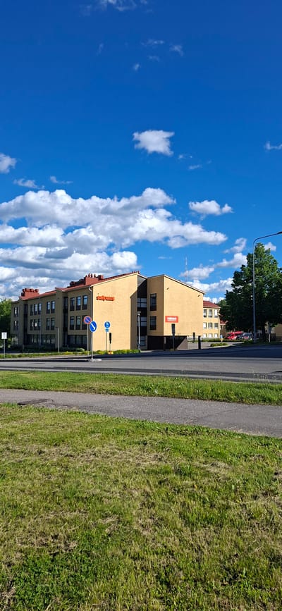 The image shows a street view in Lahti, Finland, on a clear day with scattered clouds. A light yellow building with a red roof is prominently featured. The building has multiple windows and appears to be an office or commercial space, with "Salpaus" branding visible in red letters on its facade. There are traffic signs, including a blue sign with a bicycle symbol and a white sign with a blue circle and a red diagonal line, indicating traffic regulations. A paved road runs in front of the building, bordered by a grassy area in the foreground. Trees and other buildings are visible in the background. The overall scene depicts an urban environment in daylight.